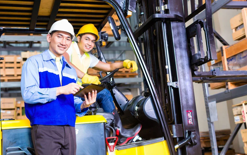 Two warehouse workers in safety helmets operating a forklift, one holding a tablet while the other sits in the driver seat beside storage racks.