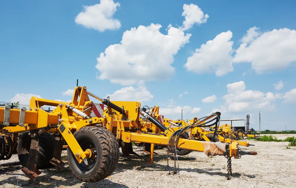 Yellow agricultural tillage equipment with large tyres and hydraulic hoses parked on a gravel lot under a blue sky.