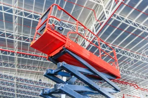 Red scissor lift platform raised inside an industrial warehouse, reaching the ceiling structure for maintenance or installation work.
