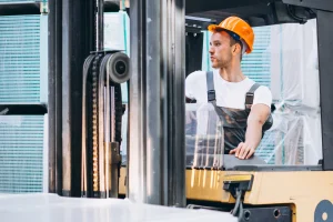 Worker in orange hard hat operating a forklift inside a warehouse, seated in the cab beside the mast and lift chains.