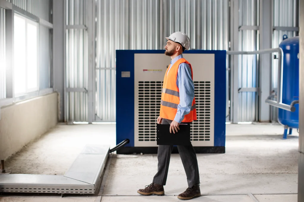 Industrial worker wearing safety helmet and vest inspecting air compressor equipment inside a factory facility